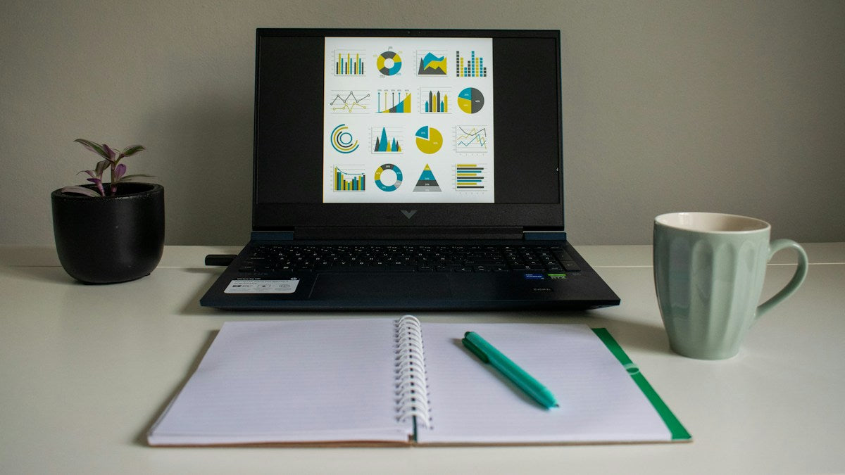 Business charts on a laptop beside a notebook for planning.
